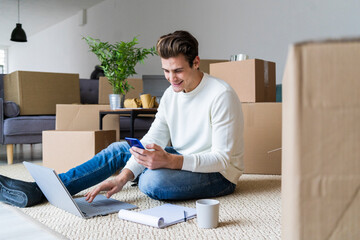 Smiling man with laptop using smart phone while sitting in living room of new home