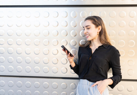 Smiling businesswoman using mobile phone while standing with hands in pockets against silver wall