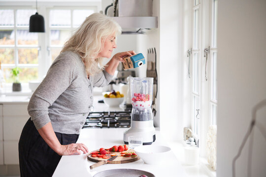 Senior Woman Pouring Milk In Processor While Preparing Strawberry Smoothie At Kitchen