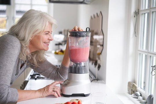 Smiling retired woman preparing strawberry smoothie in processor at kitchen counter