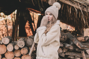 
A beautiful young girl of model appearance dressed in winter clothes holds hands in mittens near her face on the background of firewood