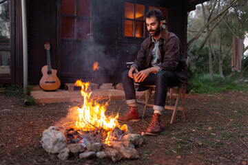 Young man with coffee cup sitting on chair near bonfire by wooden house