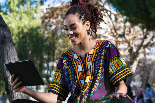 Smiling beautiful Jamaican woman with dreadlocks using digital tablet at park on sunny day