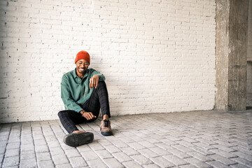 Happy man in knit hat sitting on floor against white brick wall