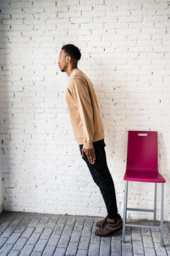 Man Leaning By Chair Against White Brick Wall At Home