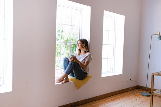 Woman with coffee cup sitting on window sill at home