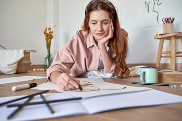 Female drawing artist with hand on chin working on drawing while lying in living room