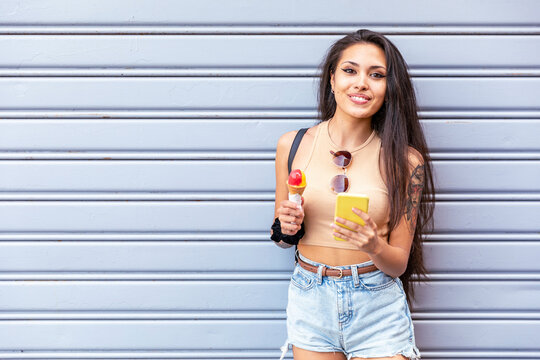 Smiling woman with ice cream cone using mobile phone while standing against shutter