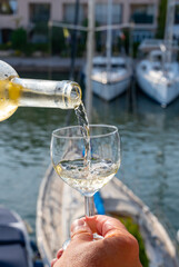 Pouring of cold white wine in glass in summer with sail boats haven of Port Grimaud on background, Provence, France