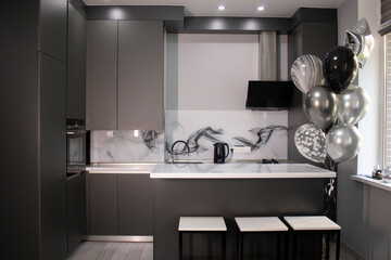 Corner of stylish Minimalistic grey kitchen, dark gray cupboards, white countertops and bar with stools. black sink, kettle and stove. and black and white balloons.