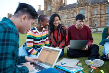 Group of young university students hanging out sitting on grass studying and using devices