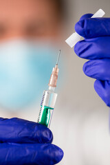 A doctor wearing a blue surgical face mask and blue gloves holding a disposable syringe with transparent injection solution in one hand and its cap in another; face is blurred; portrait orientation