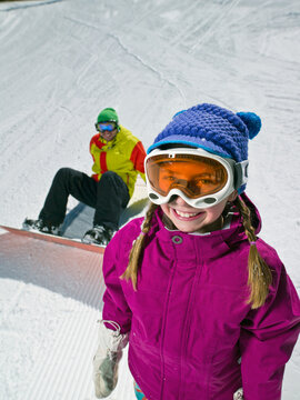 Daughter (10-11) posing with father sitting on ground with snowboard in background
