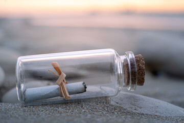 close-up view of a small glass bottle containing a paper message. The bottle is lying on the beach at sunrise.