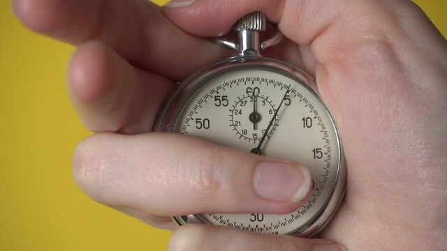 A woman's hand holds an analog stopwatch on a yellow chrome screen. The time starts with the old chronometer, the person presses the Start button in the sports concept. The concept of time management.