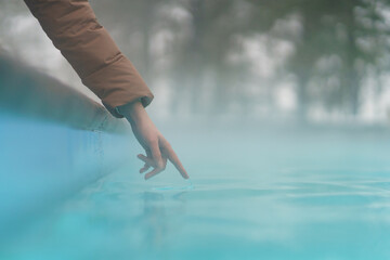 Hand of woman checking temperature of water in open swimming pool in winter. Thermal spa outdoors. 