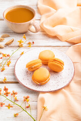 Orange macarons or macaroons cakes with cup of apricot juice on a white wooden background. Side view.