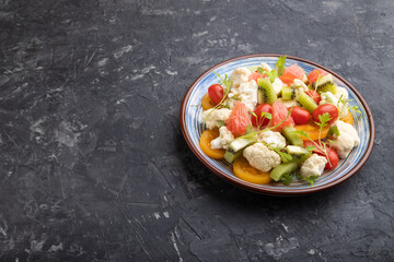 Vegetarian salad of cauliflower cabbage, kiwi, tomatoes, microgreen sprouts on black concrete background. Side view, copy space.