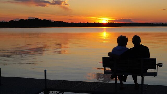 Silhouette of two people on a bench on a dock as they enjoy a sunset over a beautiful lake in Minnesota on a serene and relaxing evening, in golden hour light.