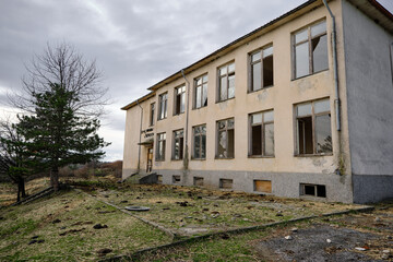 Fototapeta na wymiar 07.01.2021. Bulgaria, Kardzali. Old brownfield and abandoned soviet type school with overcast and very cloudy sky. School inside the green grass with broken windows.