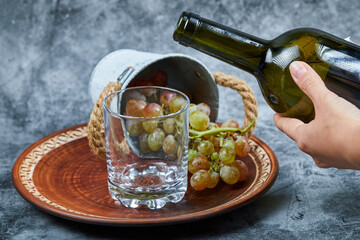 Small bucket of grapes inside ceramic plate and hand pouring wine into the glass on a marble background