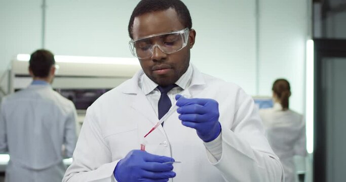Close up of happy African American young man laboratory worker in goggles works in modern lab in clinic pouring blood sample on glass conducting experiment, analysis concept, genetics, microbiology