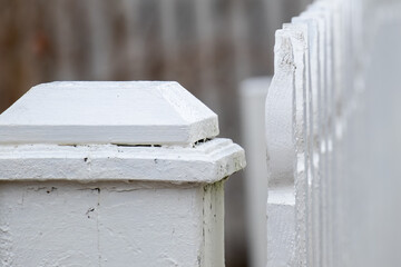 A close up of an antique white wooden picket fence post with vertical wooden pailings having a curved shape on top. The top of the country barricade post has a square wood cap. The paint is worn.