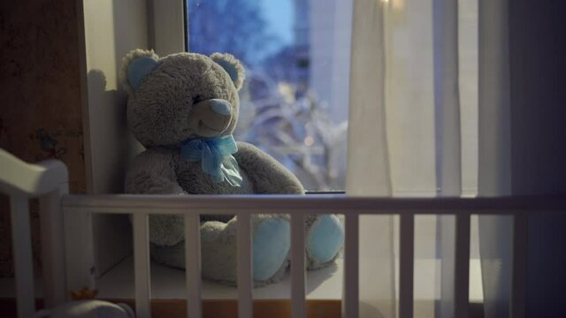 white baby cot bed soft and toy teddy bear in front of falling snow outside the window at peaceful winter night