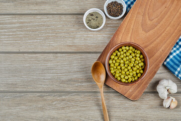 A bowl of boiled green peas with a spoon, spices, garlic, and a blue tablecloth on a wooden table