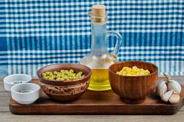 Bowls of boiled sweet corn and green peas, spices, oil, and vegetables on a wooden board with a tablecloth