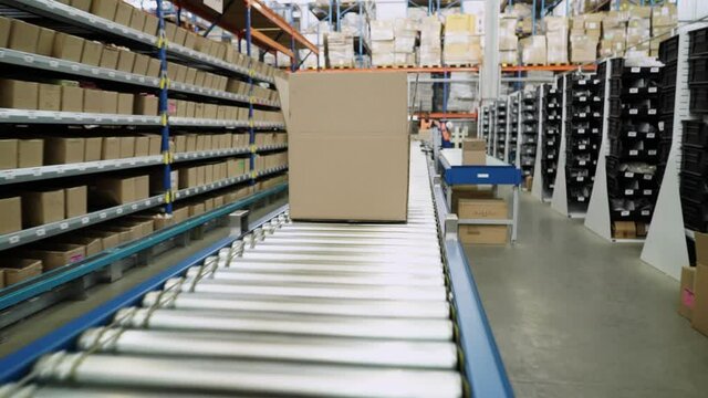Camera tracks cardboard package on automated roller conveyor in large distribution warehouse.