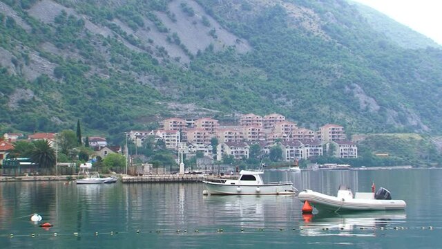 Fishing boat and dinghy anchored in the water. Coastal town with buildings and mountains in back. Boka Kotorska Bay in Montenegro.
