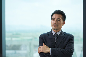 contemplative asian business man standing by windwo in office - obrazy, fototapety, plakaty