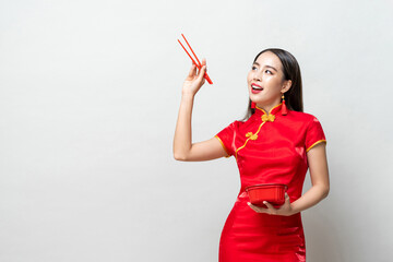 Asian woman in traditional red Chinese qipao dress holding plastic food box and chopsticks looking upward to empty space in studio isolated light gray background