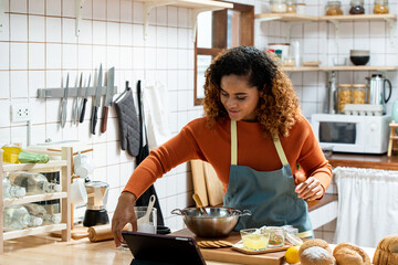 Happy young African American woman cooking in kitchen follwing online recipe in digital tablet,...