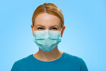 Charismatic , happy woman with surgical mask on , smiling under the mask . Medical personnel or civilian on blue background 