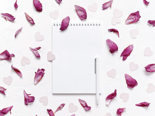 Open blank notebook, pen, peony petals, heart-shaped confetti are on a textured background. Floral layout for Valentine's day. Flat lay. Top view. Copy space.