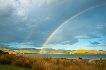 Fototapeta na wymiar Sun strikes rolling farmland across the bay arched under rainbow under dark cloudy sky