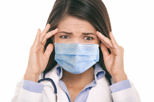 Doctor wearing coronavirus surgical face mask in panic scared doing a funny stressed out facial expression holding head from headache. Asian woman portrait.