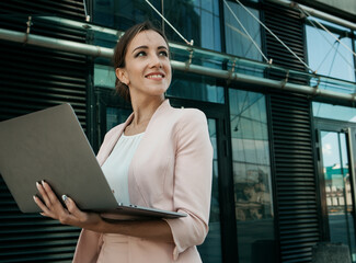 Business, communication, technology and people concept - young smiling businesswoman with laptop in the street with office buildings in the background