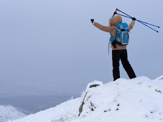 Winter hiking, joy of overcoming. Human with a backpack and nordic walking sticks in hand stands in the snow on the mountains top outdoor, selective focus.