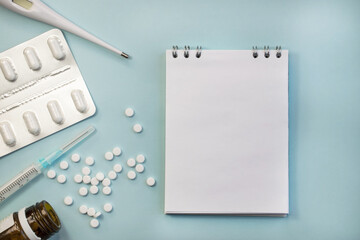 On a blue background next to the medicines, there is a notebook with a clean white sheet for the text. Thermometer and tablets on a blue background. The concept of treating colds viruses and flu .