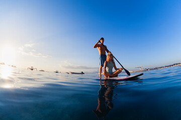 Young happy couple have fun on stand up paddleboard. Active paddle boarder paddling by sunset sea....