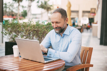 Adult male businessman sitting on the street in a cafe, drinking coffee and working on a laptop