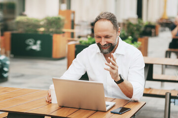 An adult man sits on the street in a cafe, communicates on the network and works on a laptop