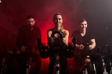 Fototapeta premium fit people riding exercise bikes in gym, enjoying workout time, in smoky red neon space