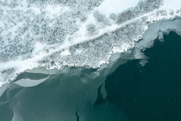 aerial top view of frozen river with cracked ice and snow-covered shore during heavy snowfall