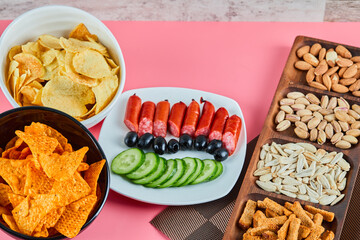 Assorted snacks on the pink table. Chips, sausages, pistachios, olives