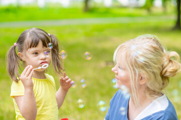Little girl with syndrome down blows bubbles in a summer park with her mother