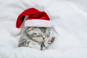 Cute kitten wearing red santa's hat sleeps under a white blanket on a bed. Top down view. Empty...